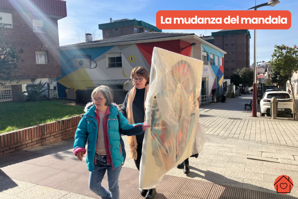 Maleni lleva el mandala por la calle con la ayuda de su hermana y un trabajador de Igualarte. La luz le ilumina. El mandala está cubierto por un plástico, pero se puede ver su colorido. De fondo el edificio de la Fundación Igualarte.